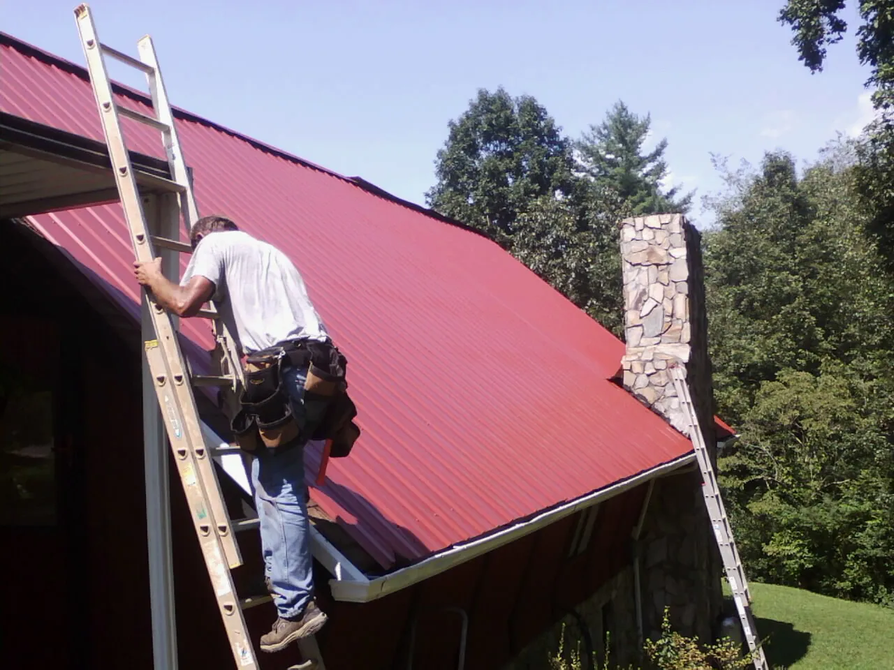Roofer on a ladder beside a red metal roof and stone chimney — Soffit & Fascia Repair by Roofing Brothers in Strasburg